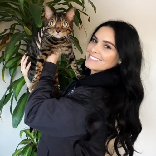 A woman with long, wavy dark hair smiles while holding a Bengal cat, standing in front of a large green plant.