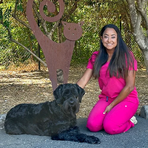 Dr. Margaret Basurto, wearing pink scrubs, kneels beside a large black dog outdoors, with a metal cat sculpture in the background.