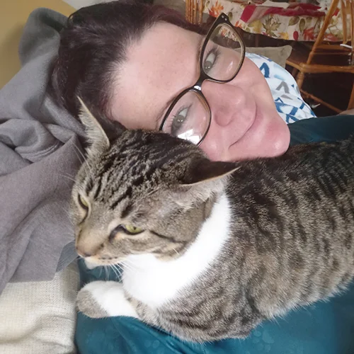 Heidi, wearing glasses, smiles while lying on a couch with a tabby cat resting on her chest.