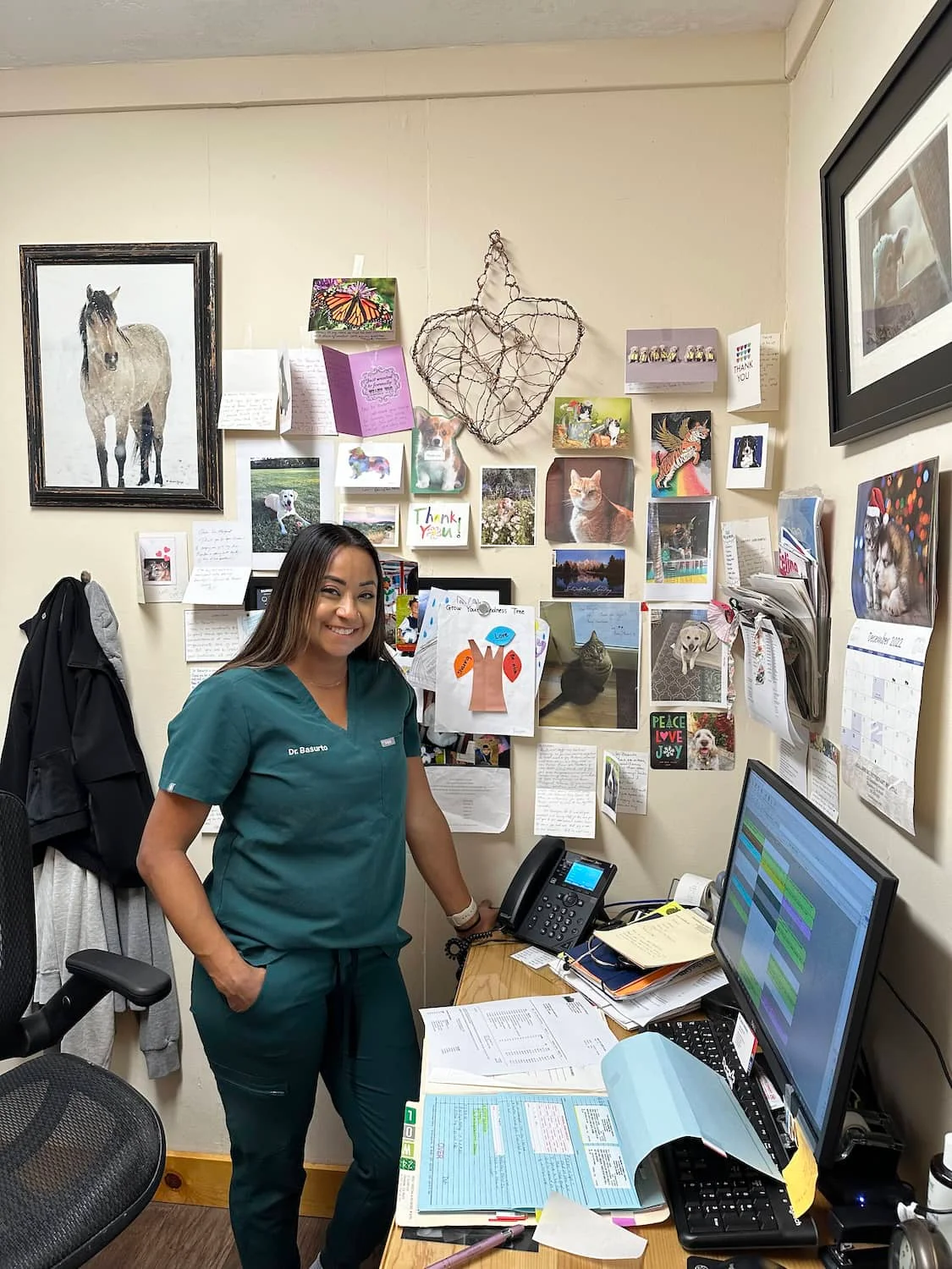 A veterinary professional in scrubs smiles while standing in an office decorated with thank-you cards and pet photos, next to a desk with a computer and paperwork.